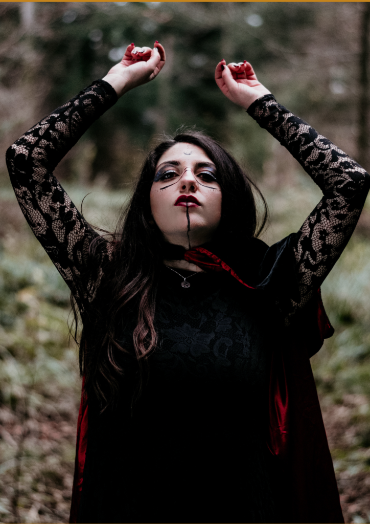 Woman with intricate makeup and black lace clothing, raising her hands gracefully in a forest setting.