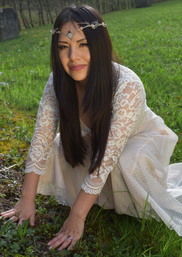 Woman dressed in white, kneeling in a field, with a tiara on her forehead.