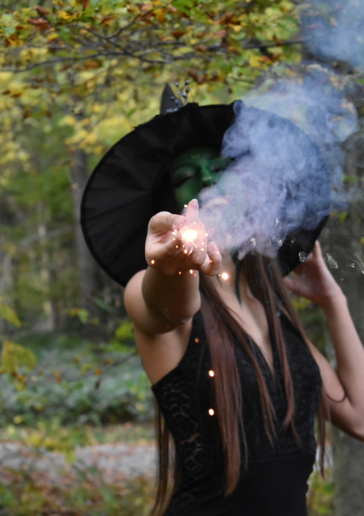 Woman with green face paint and a witch hat, extending her hand with a sparkling light and smoke swirling in the forest.