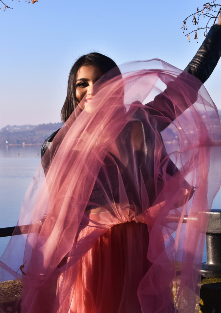 Woman in a pink gown with flowing fabric, against a lake backdrop.
