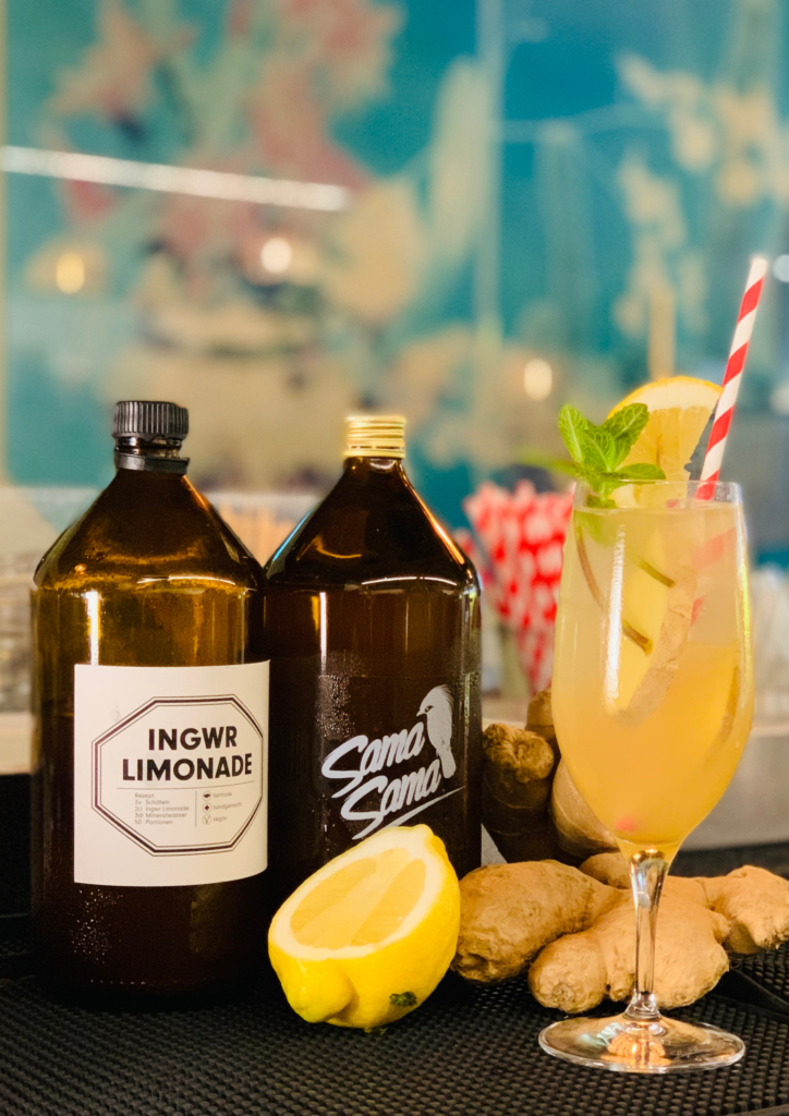 Two bottles labeled ginger lemonade beside a glass of citrus and mint-infused drink.