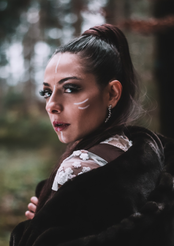 Woman with tribal makeup on her face, wrapped in a dark coat, looking back in a forest with a strong and mysterious expression.