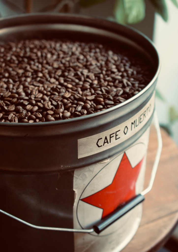 Metal bucket filled with coffee beans with a label reading “Café o Muerte.”